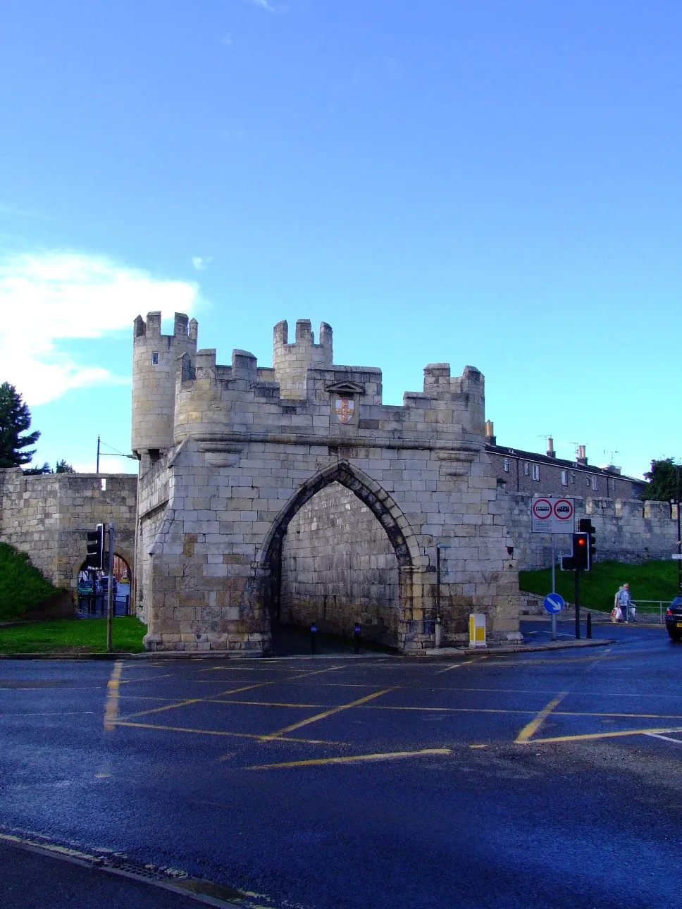 Walmgate Bar, una puerta de piedra medieval con torres almenadas y una gran entrada de arco apuntado, situada en un cruce de caminos con marcas viales amarillas bajo un cielo azul