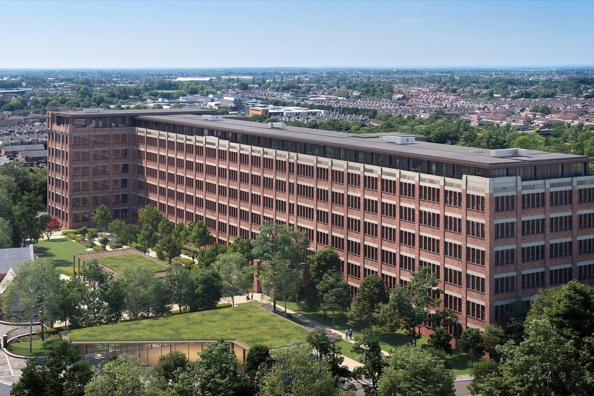 Vista aérea de un gran edificio industrial de ladrillo rojo de varias plantas con hileras de ventanas, rodeado de césped verde y árboles con el paisaje urbano de fondo