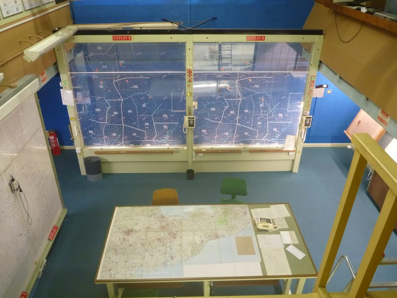 Interieur van een operatiekamer met grote verlichte wandkaarten waarop regionale routes en markeringen te zien zijn, met een tafel met een andere kaart op de voorgrond en blauwe muren.