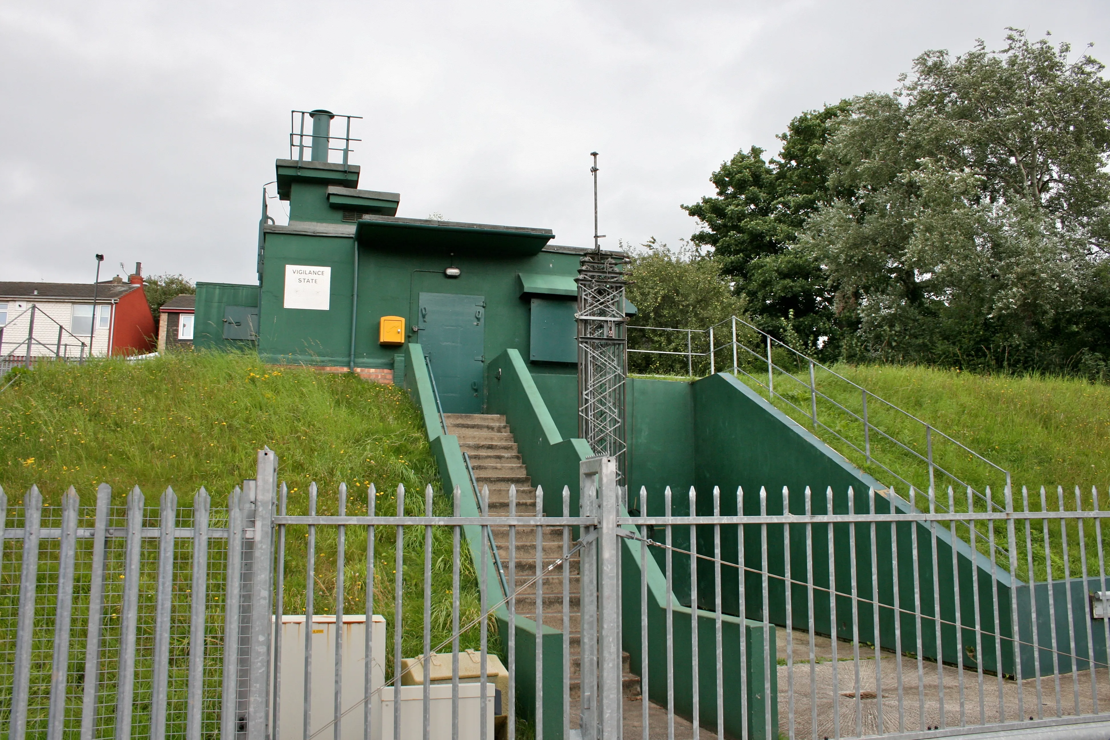Ingang van een groene betonnen bunker in een met gras begroeide heuvel, met een metalen trap die naar beneden leidt en een metalen hek rondom de locatie.