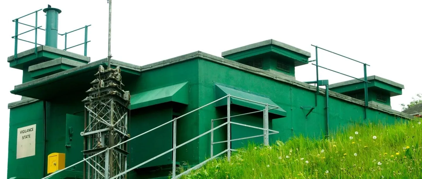 Een groen geschilderde betonnen bunker die gedeeltelijk in een met gras begroeide heuvel is ingebed, voorzien van metalen leuningen, ventilatieschachten en een bord aan de zijkant.