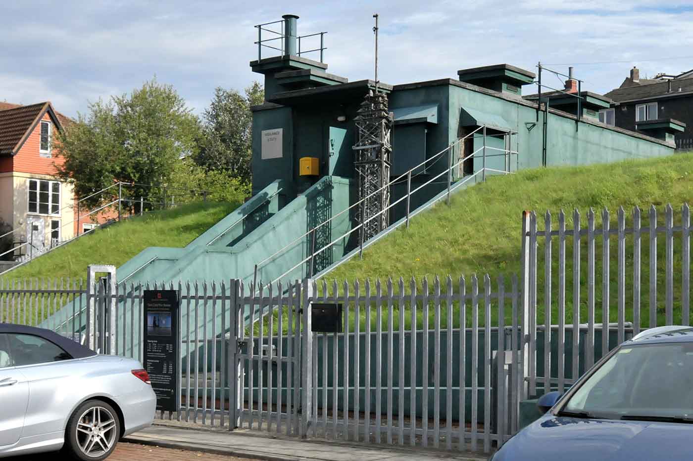 Groene, half ondergrondse betonnen bunkerstructuur gebouwd in een grasheuvel, omgeven door een metalen hekwerk met auto's geparkeerd aan de voorzijde.