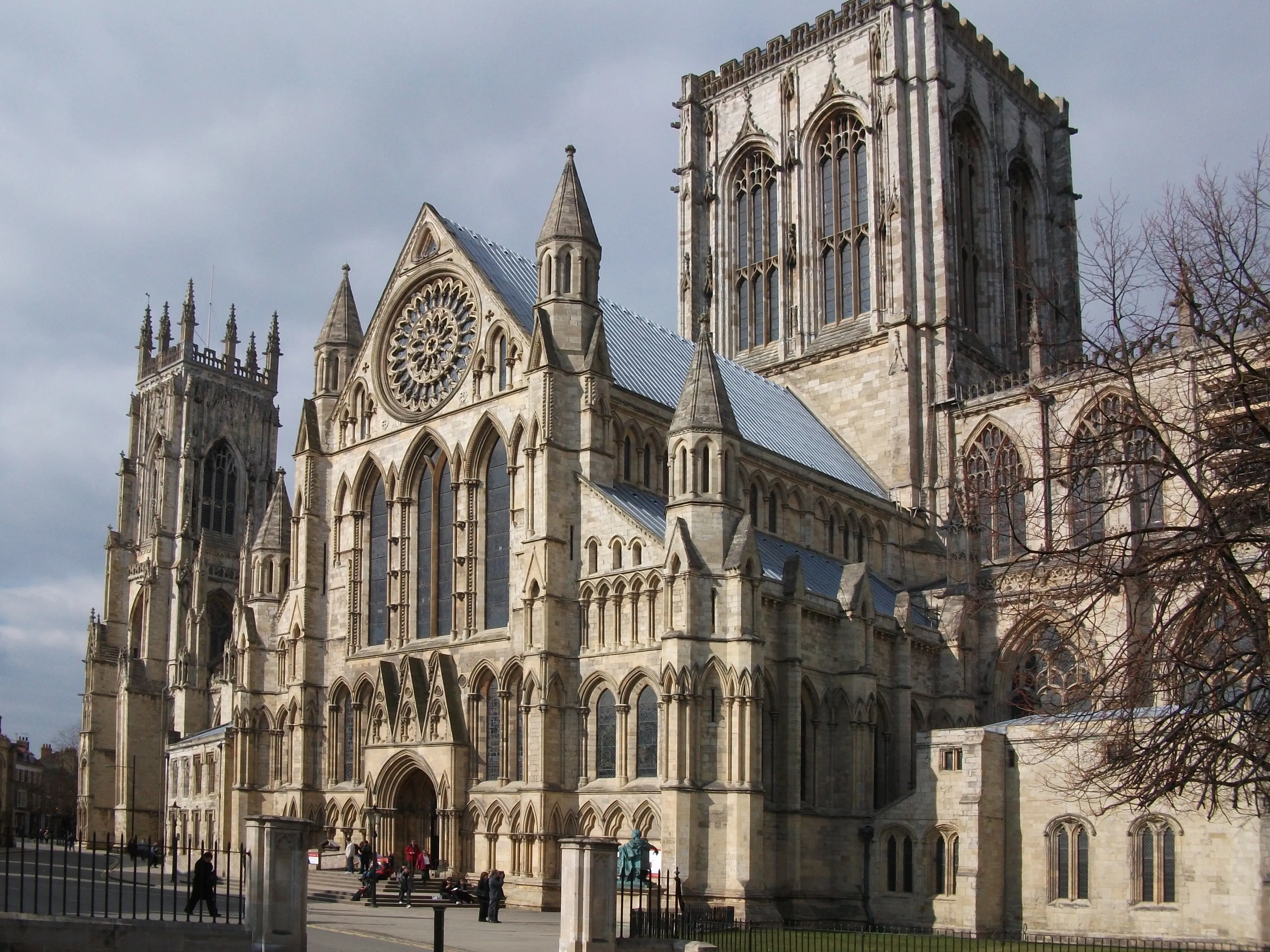 Exterior view of York Minster showing the Gothic cathedral facade with twin towers, large rose window, ornate stonework, and arched entrance under an overcast sky.