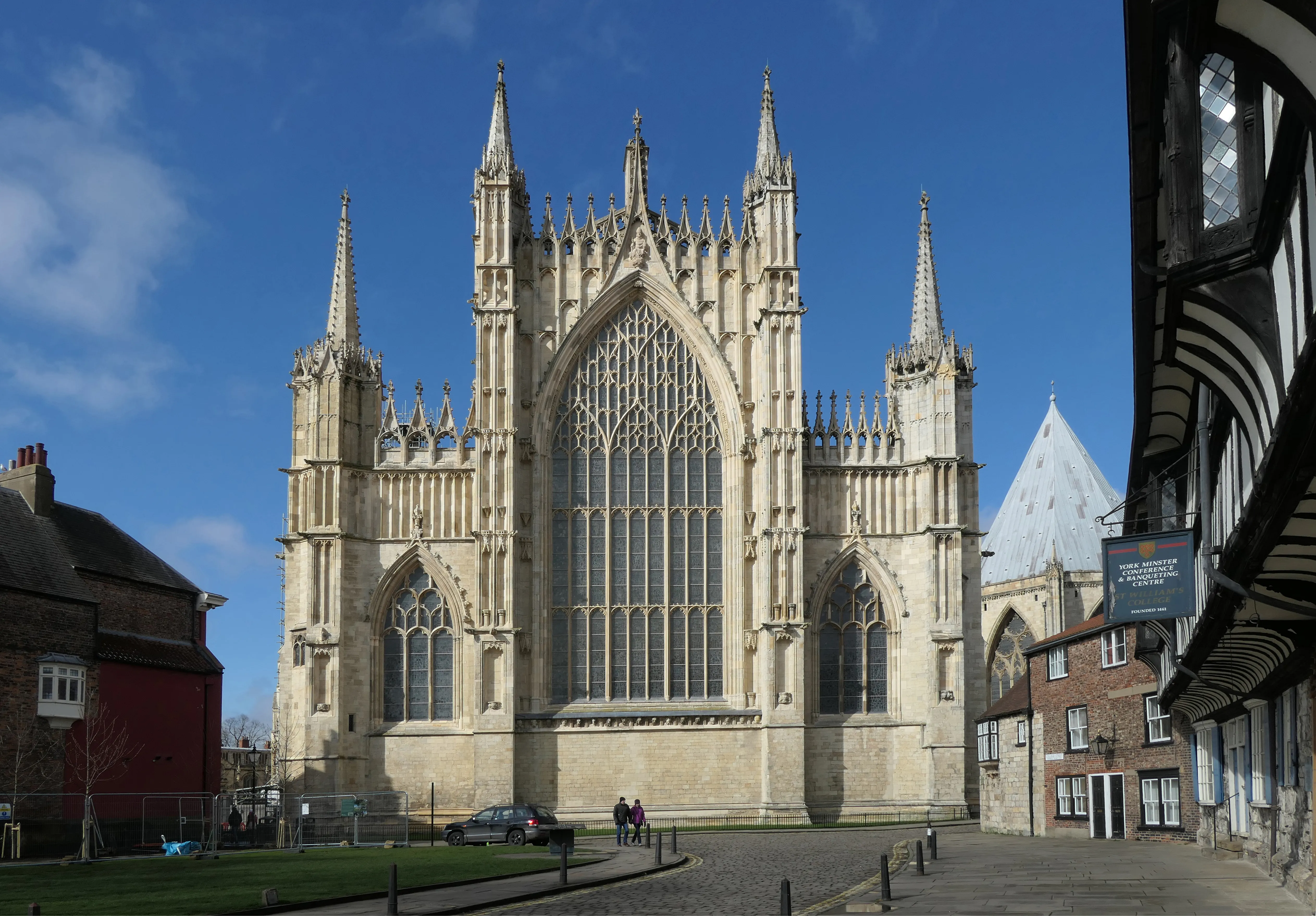 Gothic cathedral exterior with large pointed-arch stained glass window, flanking towers with pinnacles, and surrounding historic buildings under a blue sky