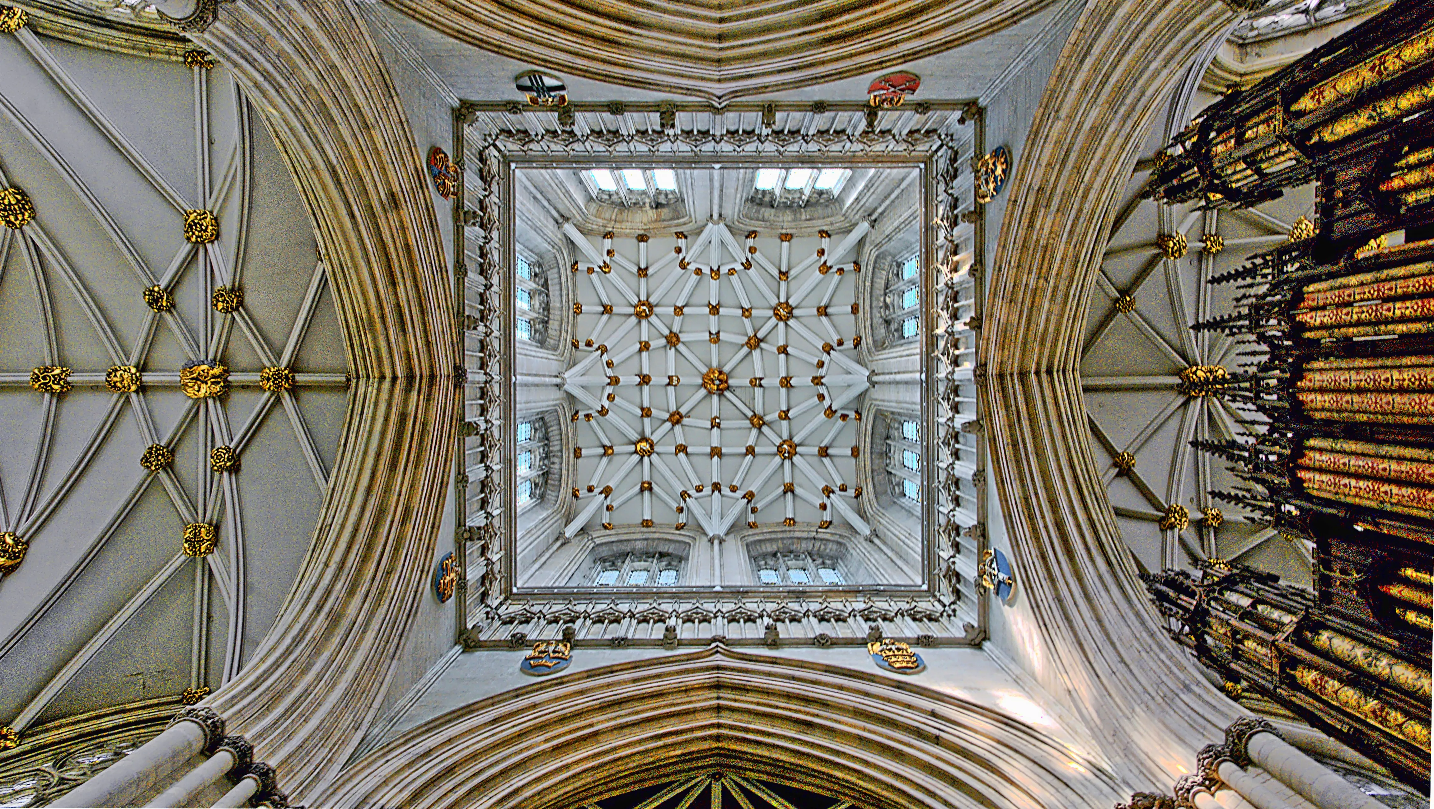 Looking straight up at the ornate ribbed vaulted ceiling of York Minster with golden decorative bosses and choir organ pipes visible on the right