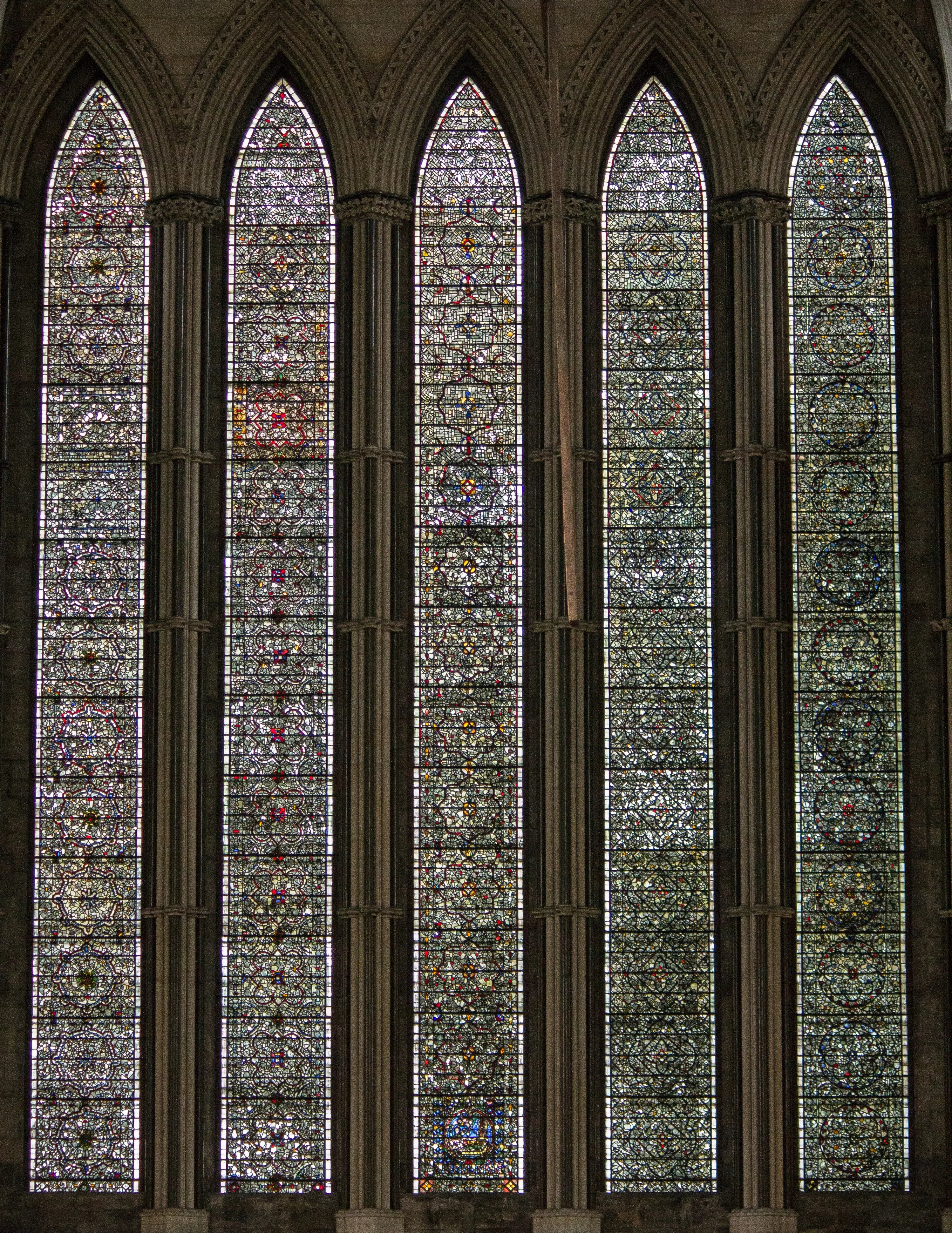 Pięć wysokich, wąskich, ostrołukowych okien witrażowych ze skomplikowanymi wzorami geometrycznymi i roślinnymi w stonowanych odcieniach zieleni, szarości oraz z domieszką czerwieni i błękitu.