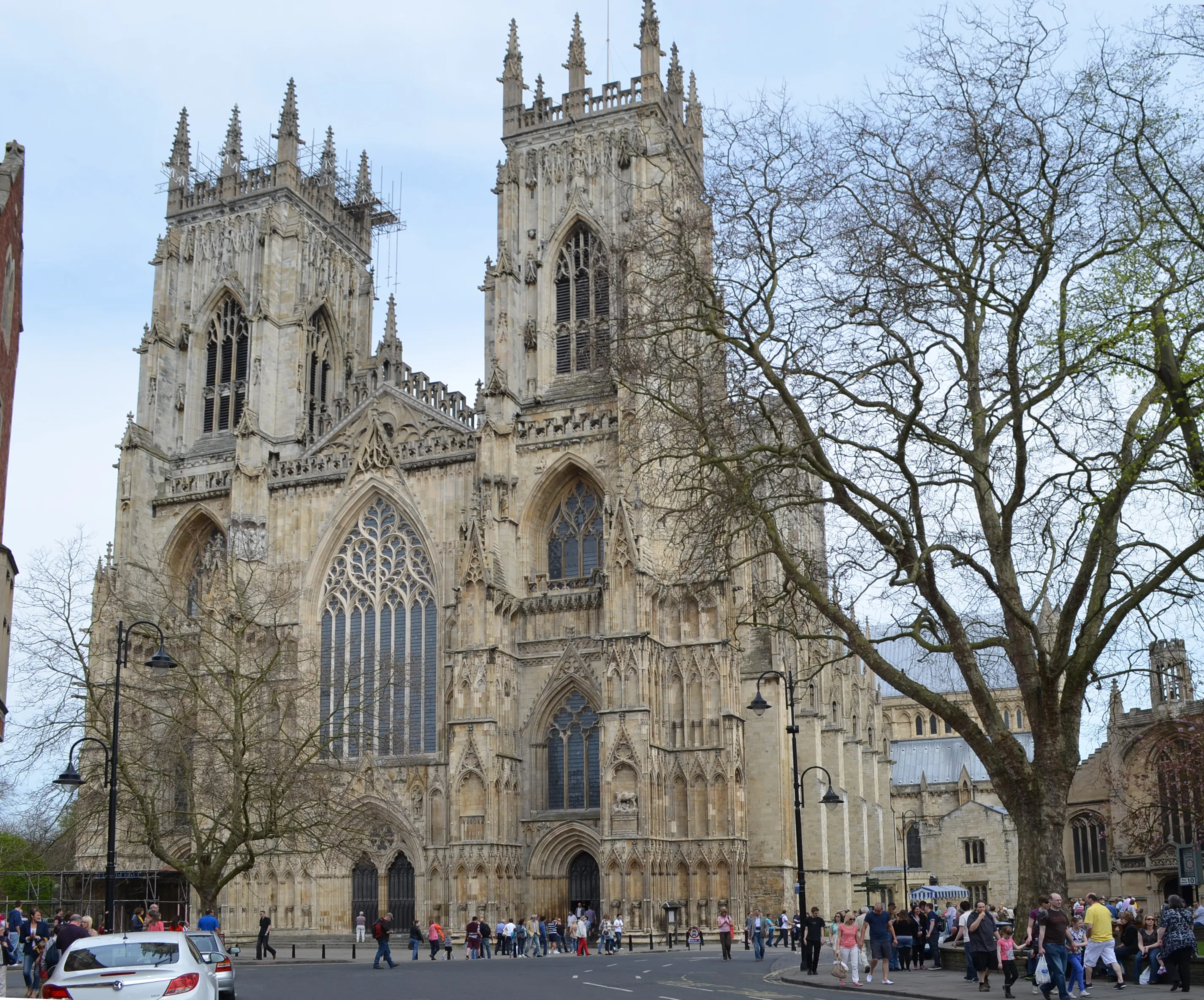 York Minster's west front facade showing two Gothic towers with pinnacles, large traceried windows, and people gathered in the plaza below