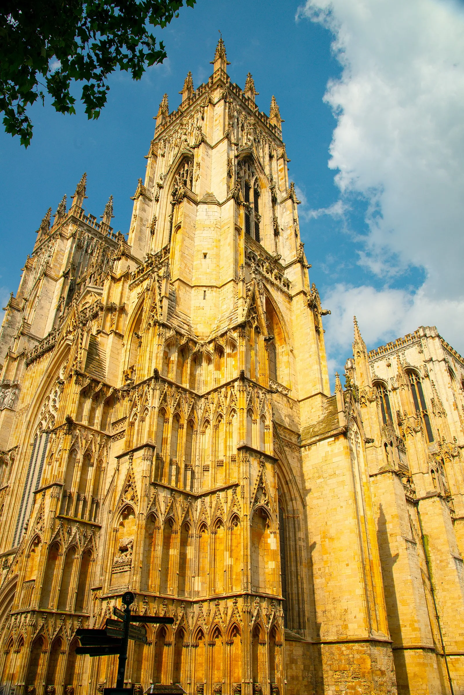 Low-angle view of York Minster's Gothic central tower and facade in warm golden stone against a blue sky with white clouds