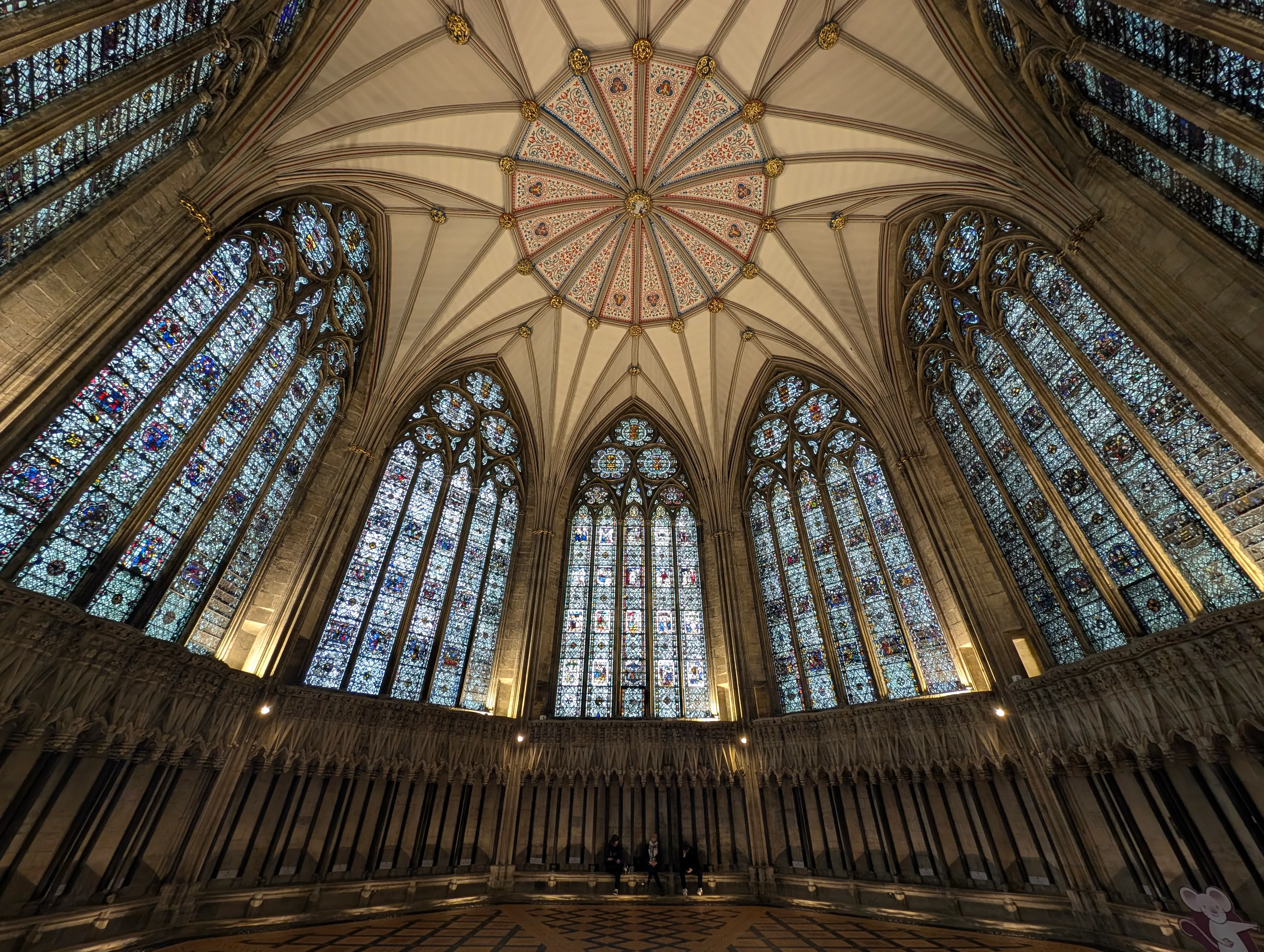 Dramatic upward view of York Minster's interior showing an ornate vaulted ceiling with a radial painted pattern and tall Gothic stained glass windows lining the walls above stone arcading.