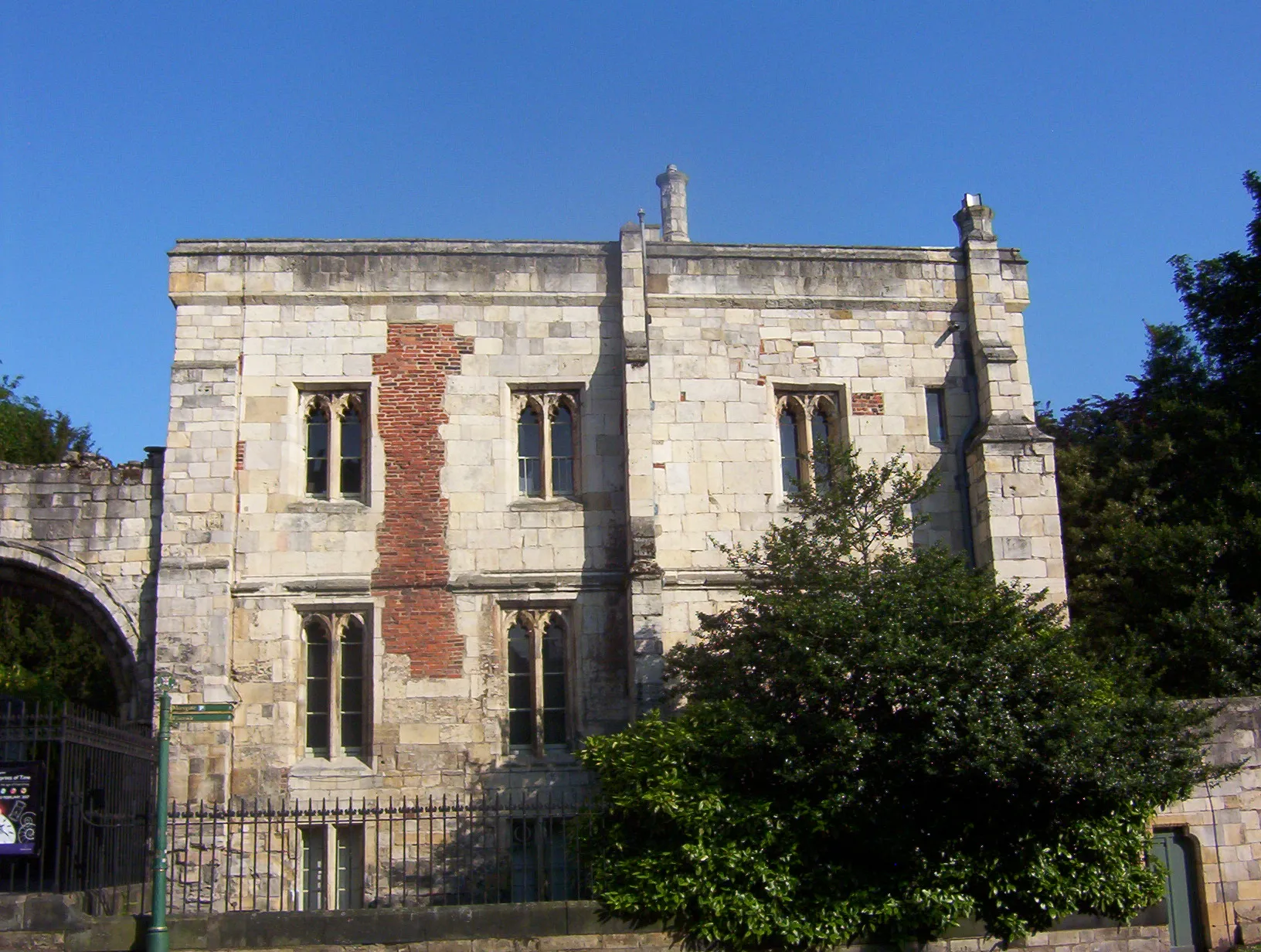 Fachada de piedra de St Mary's Lodge con ventanas arqueadas de estilo gótico, relleno de ladrillo rojo, rejas de hierro y vegetación bajo un cielo azul despejado