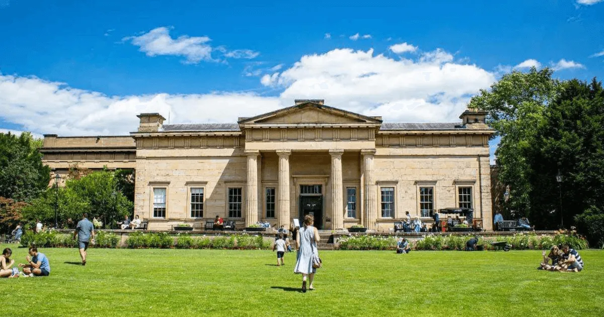 Edificio neoclásico del Yorkshire Museum con un pórtico de columnas, rodeado por un césped verde con visitantes bajo un cielo azul con nubes blancas