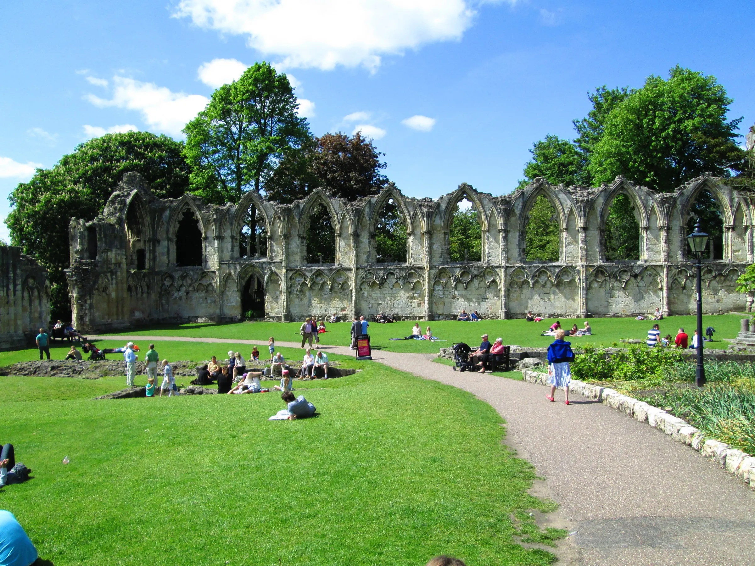 Ruinas de piedra de la abadía de St Mary's con ventanas arqueadas en los York Museum Gardens, gente descansando en el césped verde bajo un cielo azul