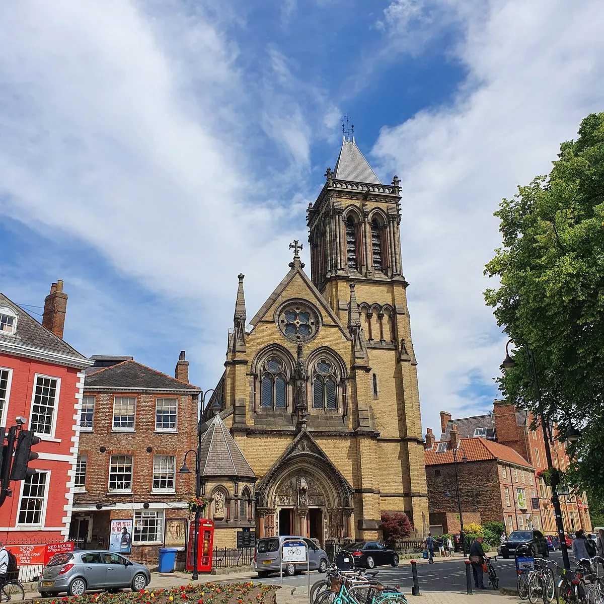 Stone church with tall bell tower and Gothic arched windows, surrounded by street with cars, bicycles, and pedestrians under blue sky with clouds