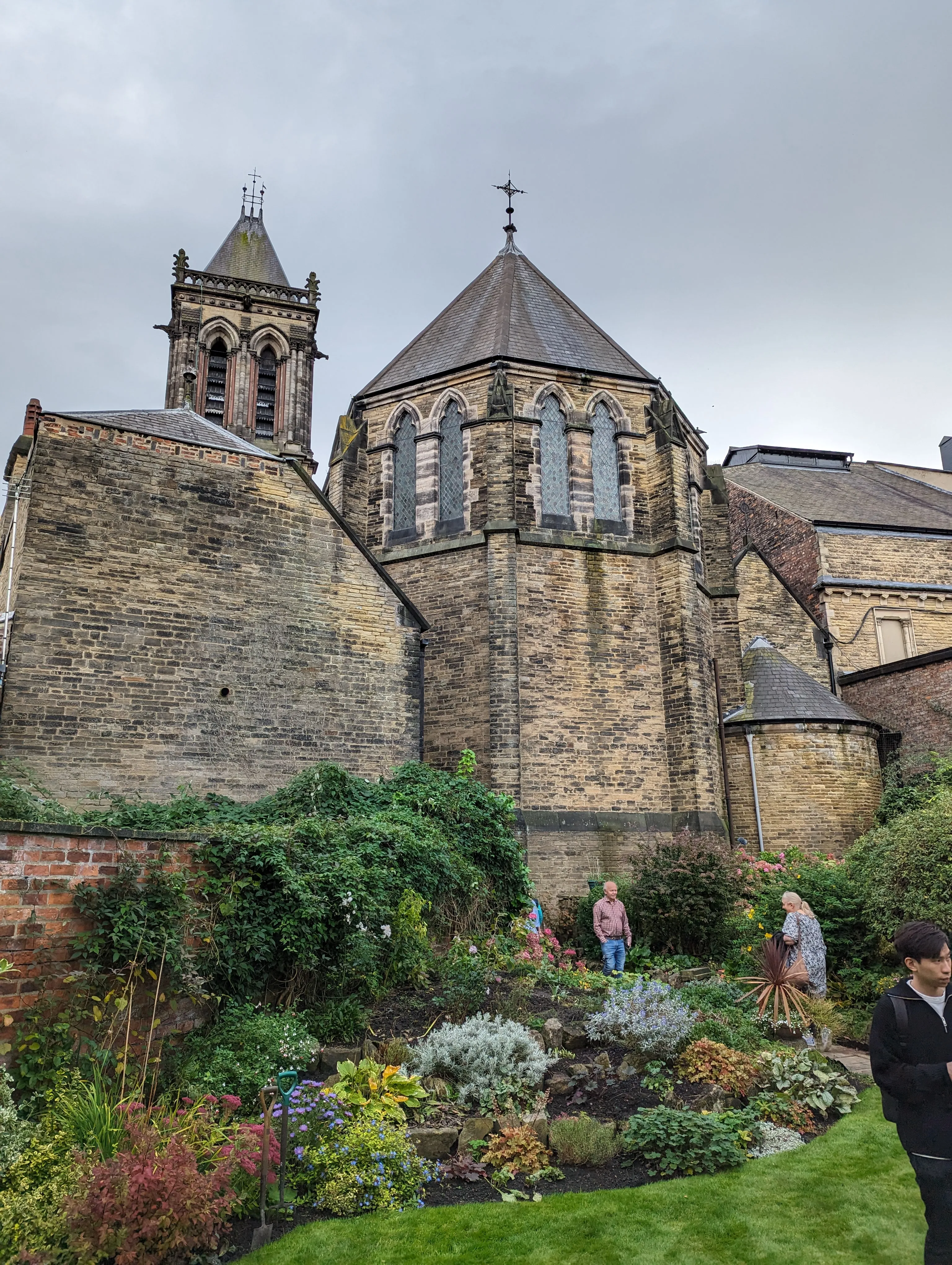 Stone church exterior showing a bell tower, octagonal apse with Gothic arched windows, and a garden with visitors in the foreground