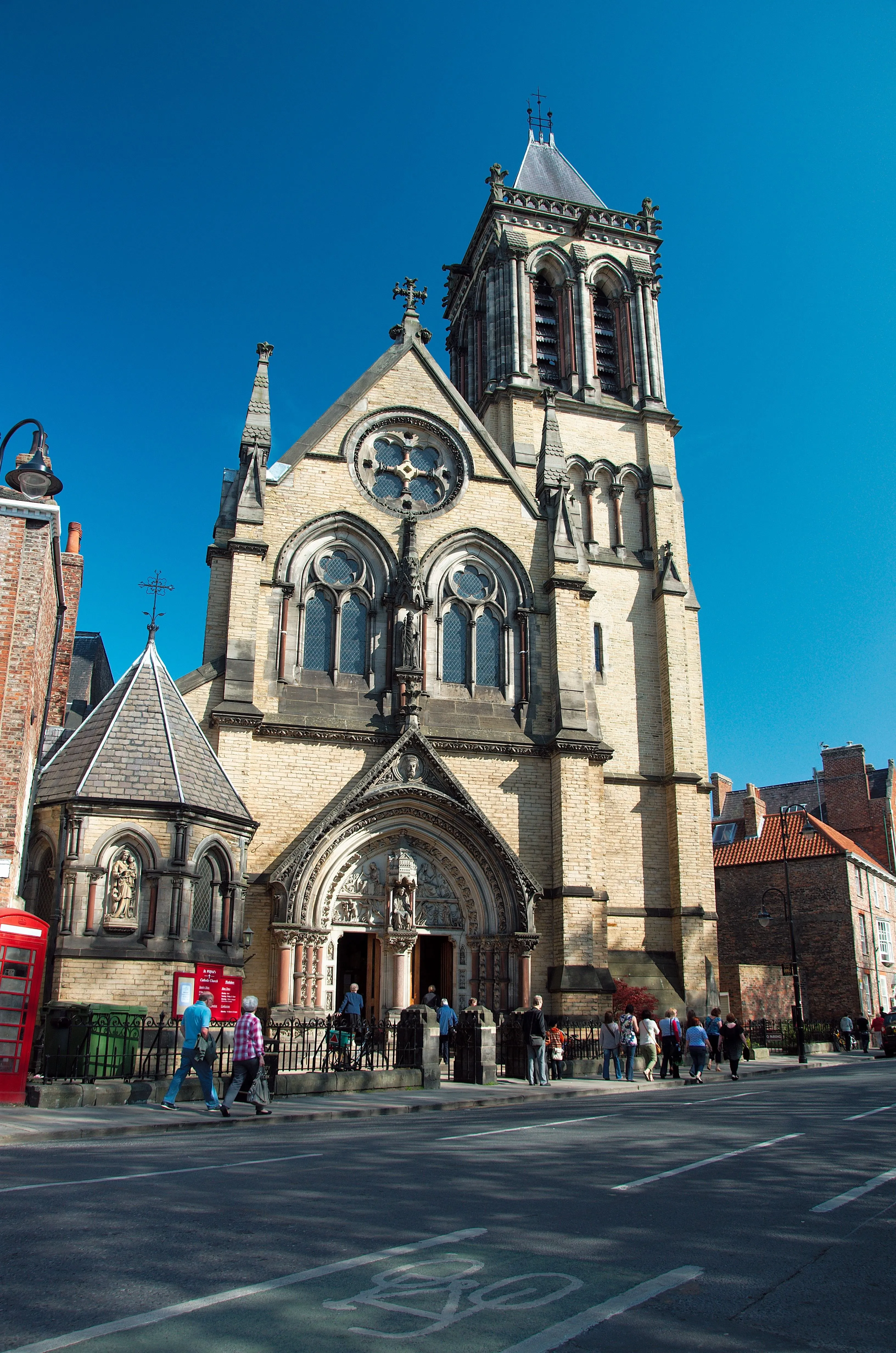 Stone Gothic Revival church facade with tall bell tower, arched windows, rose window, and people walking along the street in front under a clear blue sky.