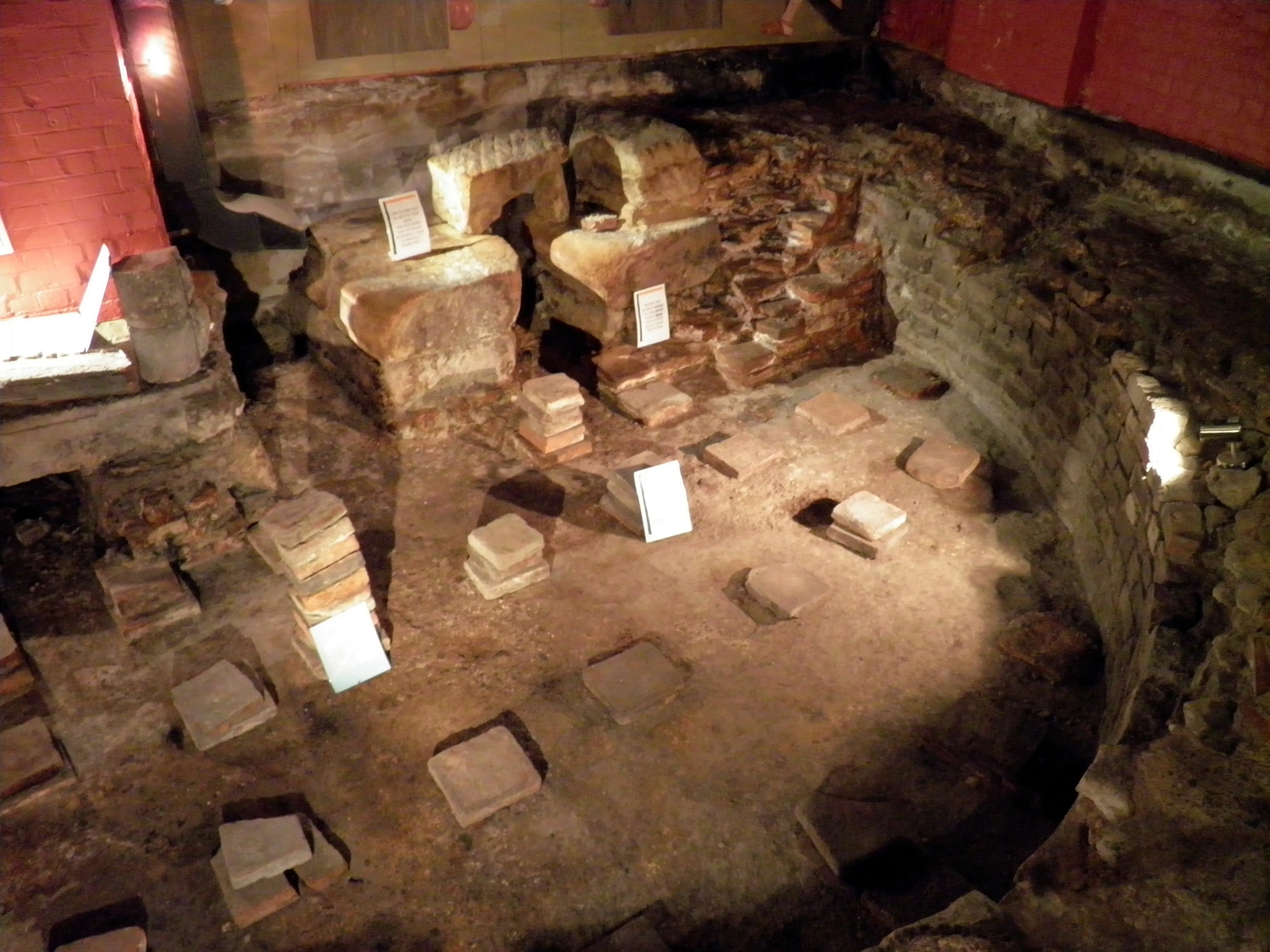 Podziemne pozostałości archeologiczne łaźni rzymskiej z ceglanymi i kamiennymi fundamentami Caldarium, z małymi białymi tabliczkami informacyjnymi umieszczonymi wśród ruin w ciepłym świetle reflektorów.