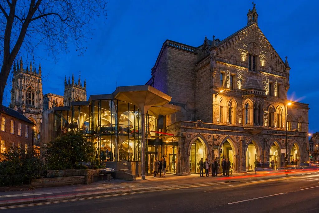 York Theatre Royal al atardecer con su fachada de piedra iluminada, su ampliación de cristal y las torres de York Minster al fondo