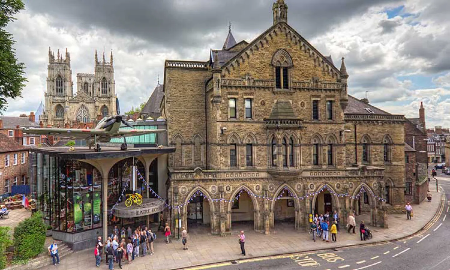 Vista exterior del York Theatre Royal, un histórico edificio de piedra con arcos góticos y una moderna ampliación de cristal en la entrada, con la York Minster visible al fondo.
