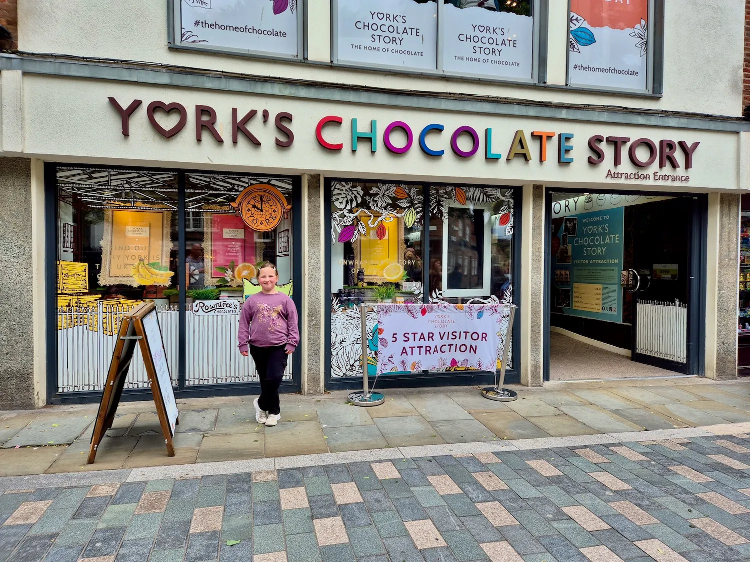Vista a nivel de calle de la fachada de York's Chocolate Story con letreros de letras coloridas sobre grandes escaparates y un visitante de pie frente a ellos en la acera