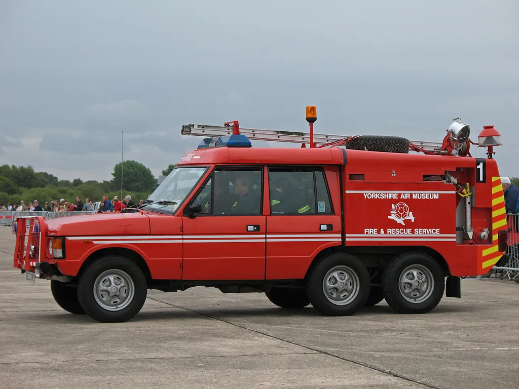Rood, zeswielig Range Rover brandweer- en reddingsvoertuig met de belettering van het Yorkshire Air Museum en een witte streep, geparkeerd op een asfaltterrein met mensen op de achtergrond.
