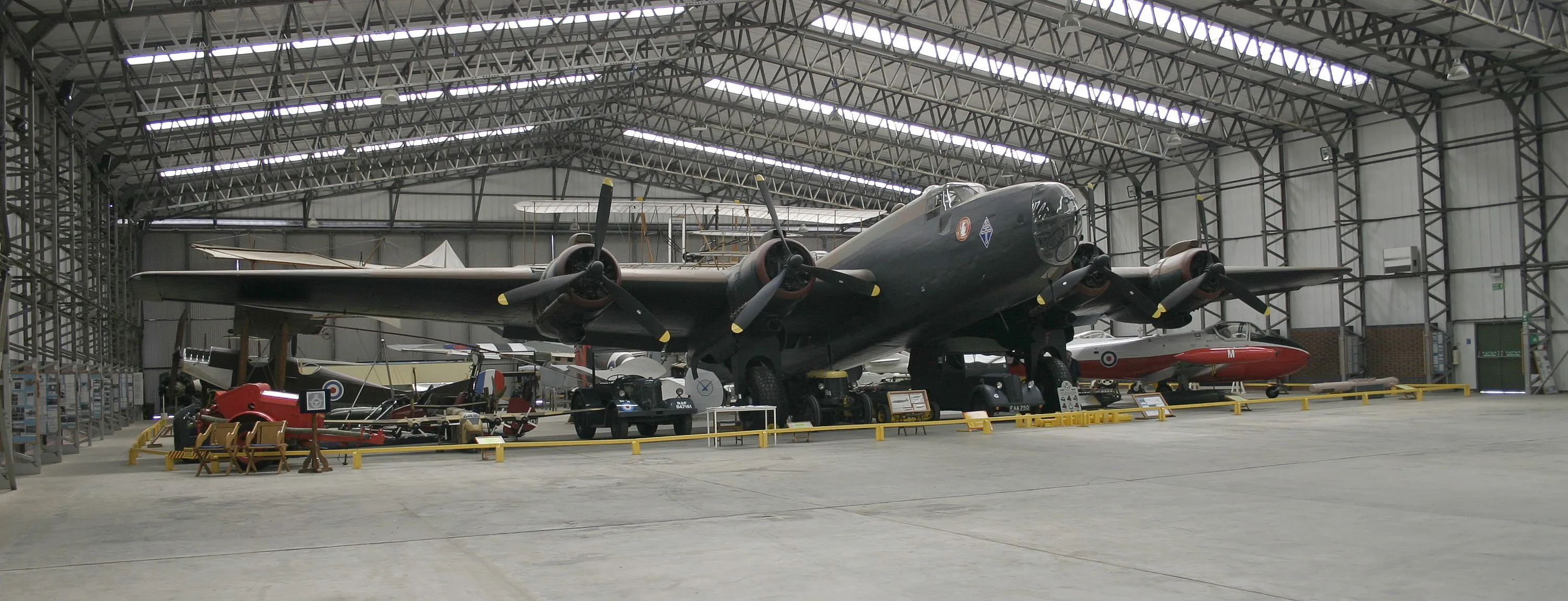 Een grote, donkere viermotorige bommenwerper staat in een ruime hangar met een hoog vakwerkplafond, geflankeerd door kleinere rode vliegtuigen en gele barrières.