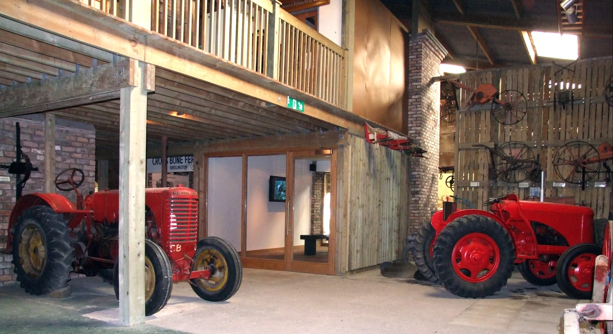 Dos tractores antiguos de color rojo expuestos en el interior de un museo que parece un granero, con un altillo de madera arriba y herramientas agrícolas montadas en la pared derecha