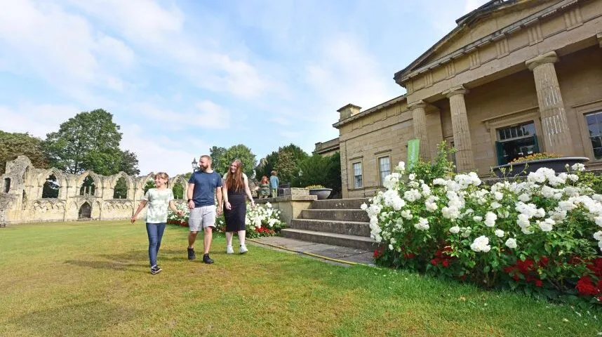 Drie mensen lopen over een groen gazon voor de klassieke gevel van het Yorkshire Museum, met witte en rode rozenstruiken en oude stenen ruïnes op de achtergrond.