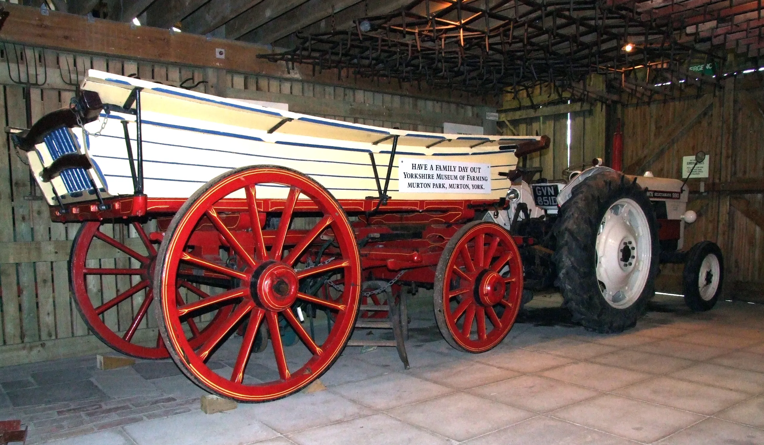 Vintage paard-en-wagen met grote rode spakenwielen en een witte houten koets, tentoongesteld naast een tractor in een museumschuur.