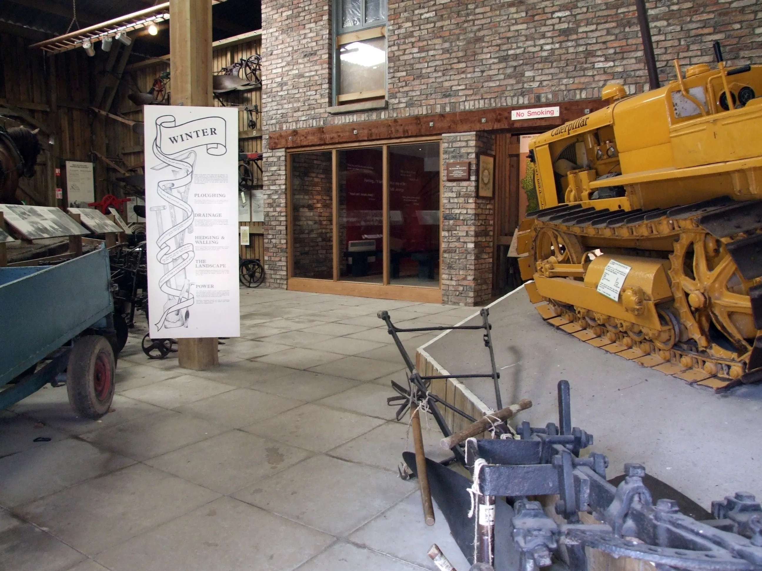 Museuminterieur met een gele rupsbulldozer en landbouwmachines, met een banner voor een wintertentoonstelling aan een houten paal tegen een bakstenen muur