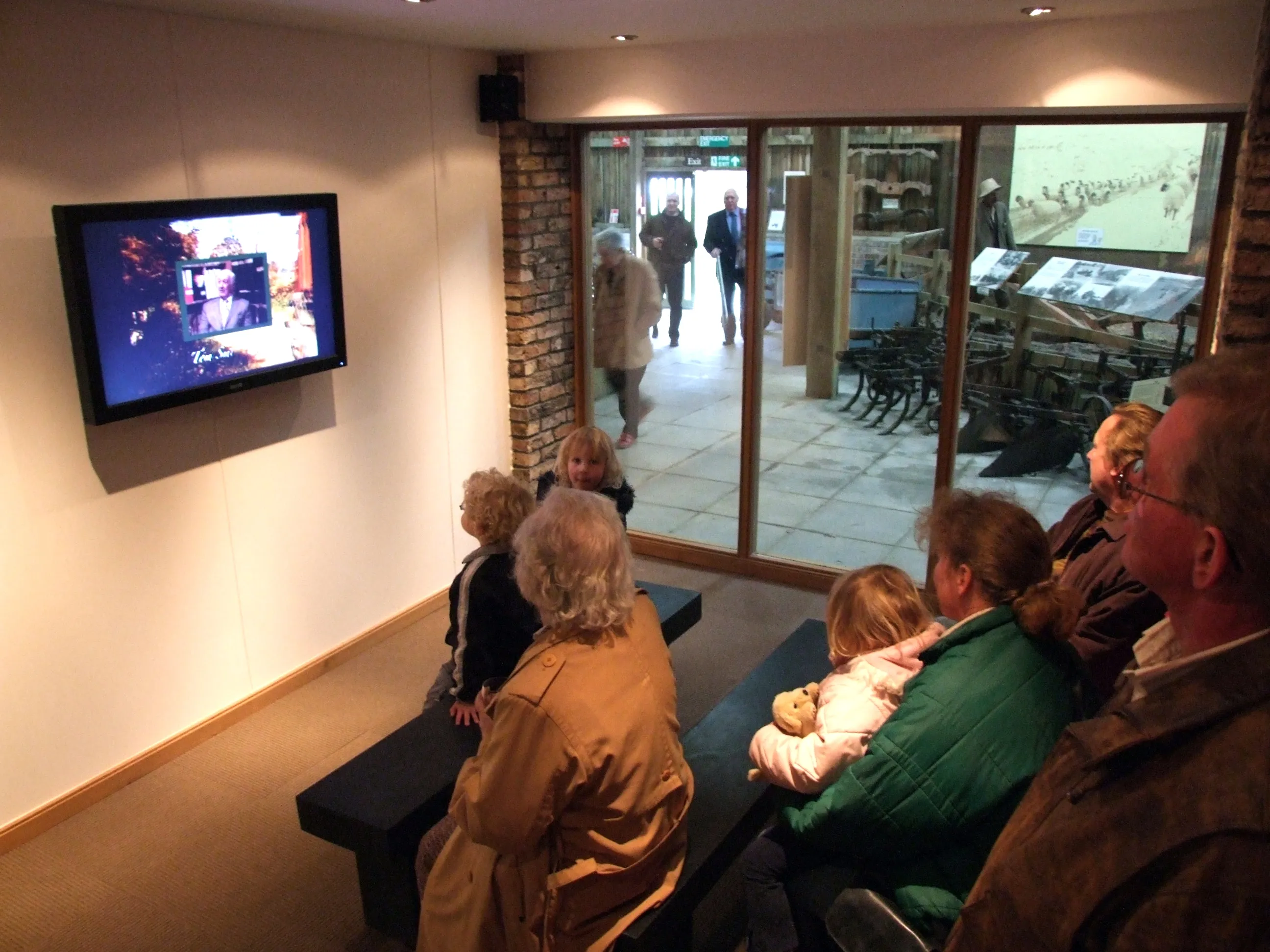 Bezoekers zitten op bankjes en kijken naar een televisie aan de muur in een museumzaal, met een groot glazen raam dat uitkijkt op een aangrenzende tentoonstellingsruimte met displays en voorbijlopende mensen.