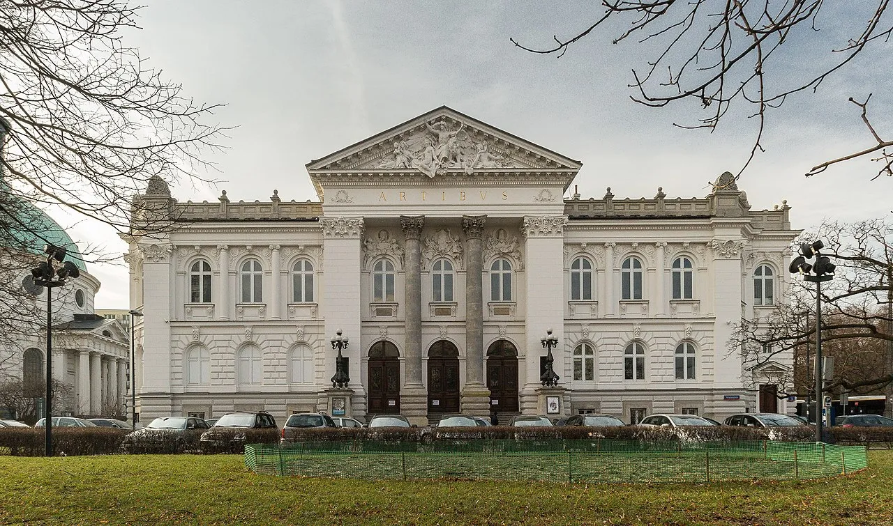 Edificio neoclásico con frontón triangular y columnas, fachada blanca, entradas arqueadas y múltiples ventanas, rodeado de árboles desnudos y un césped verde.