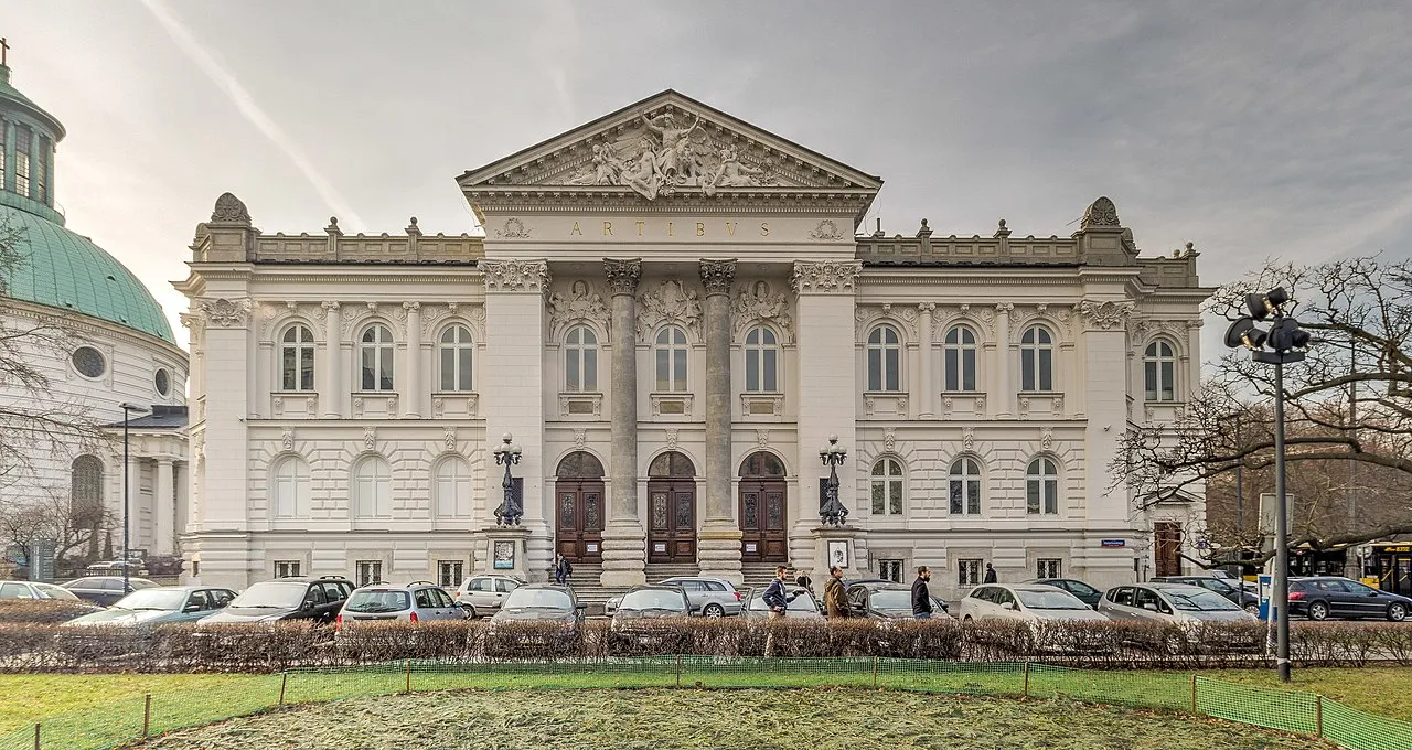 La Galería Nacional de Arte Zachęta, un gran edificio clásico con un frontón y columnas centrales, rodeado de coches aparcados y un césped verde.