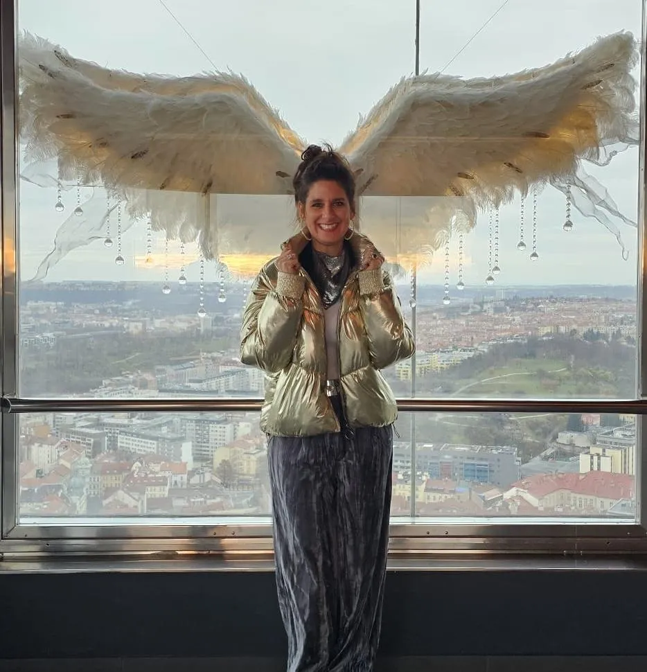 Una mujer con una chaqueta dorada está parada frente a una gran ventana con una vista panorámica de la ciudad y una gran escultura blanca alada detrás de ella.