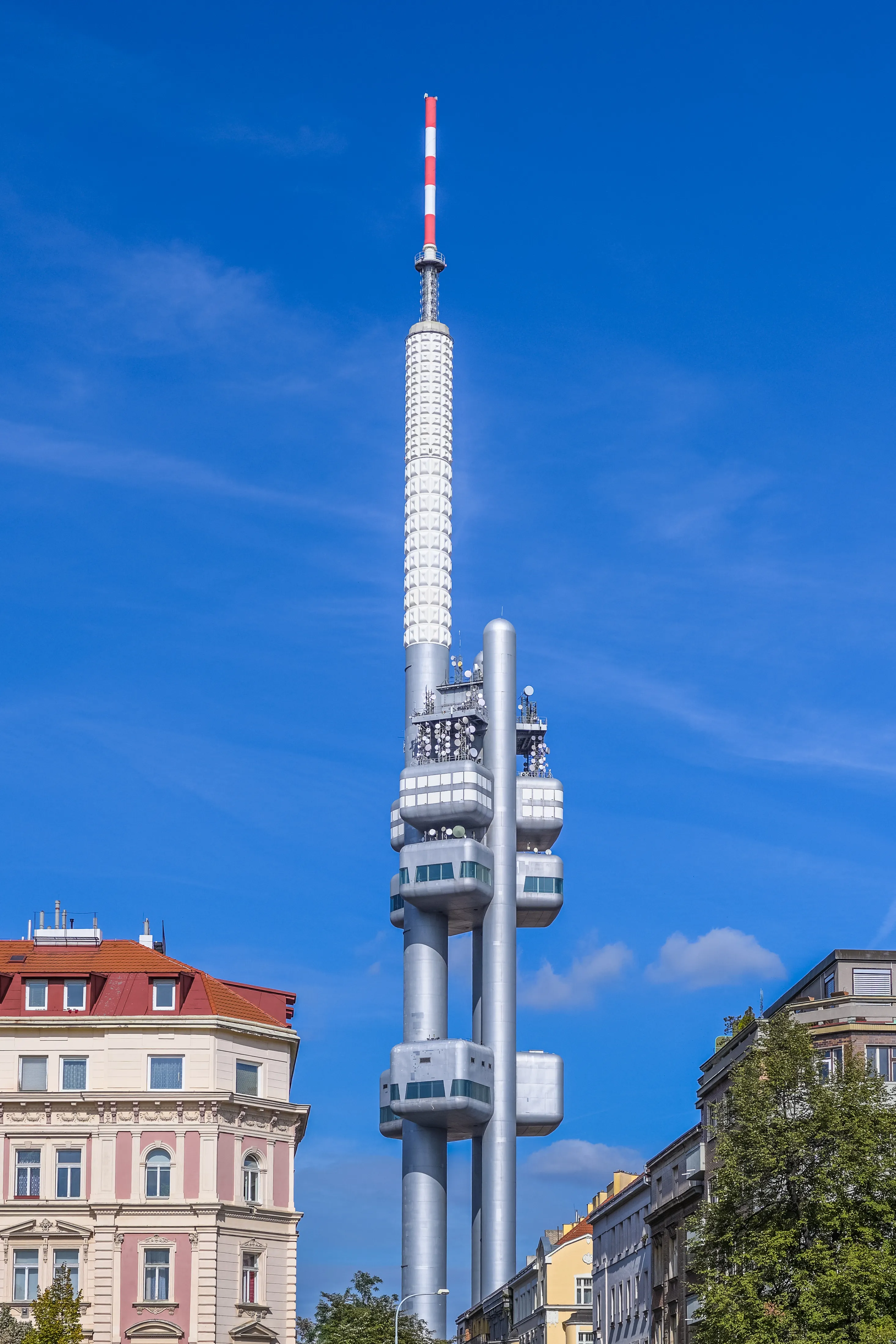 Torre de televisión plateada y alta con antena roja y blanca contra el cielo azul, rodeada de edificios