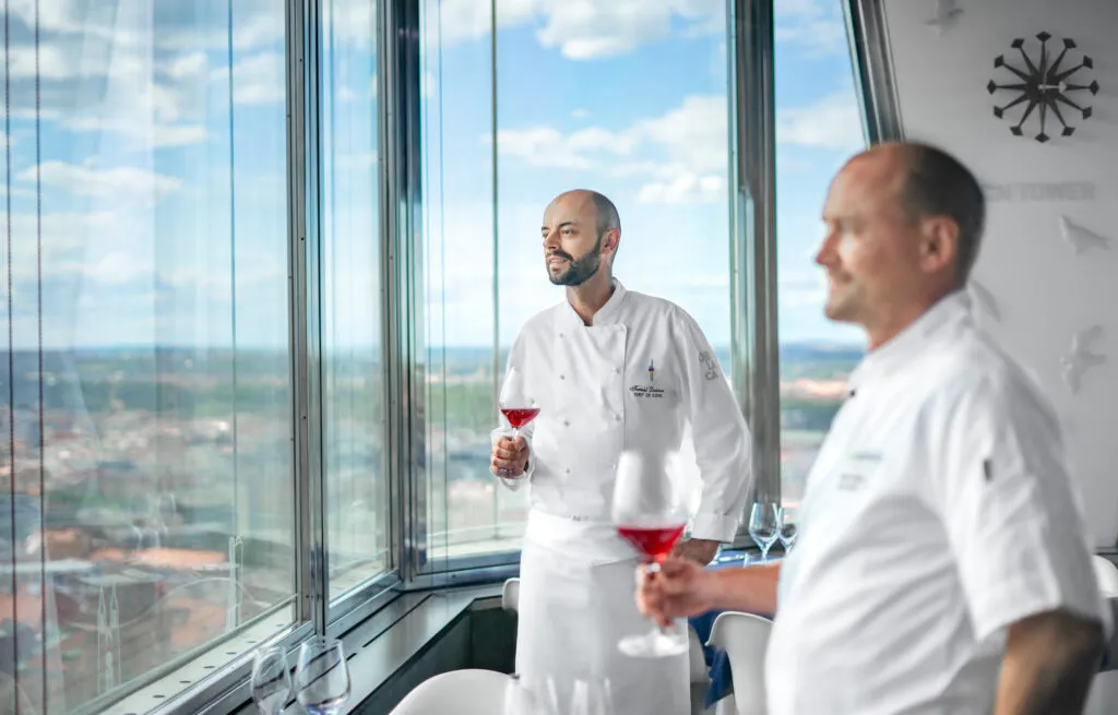 Dos chefs con uniformes blancos de pie junto a grandes ventanales con vistas a la ciudad