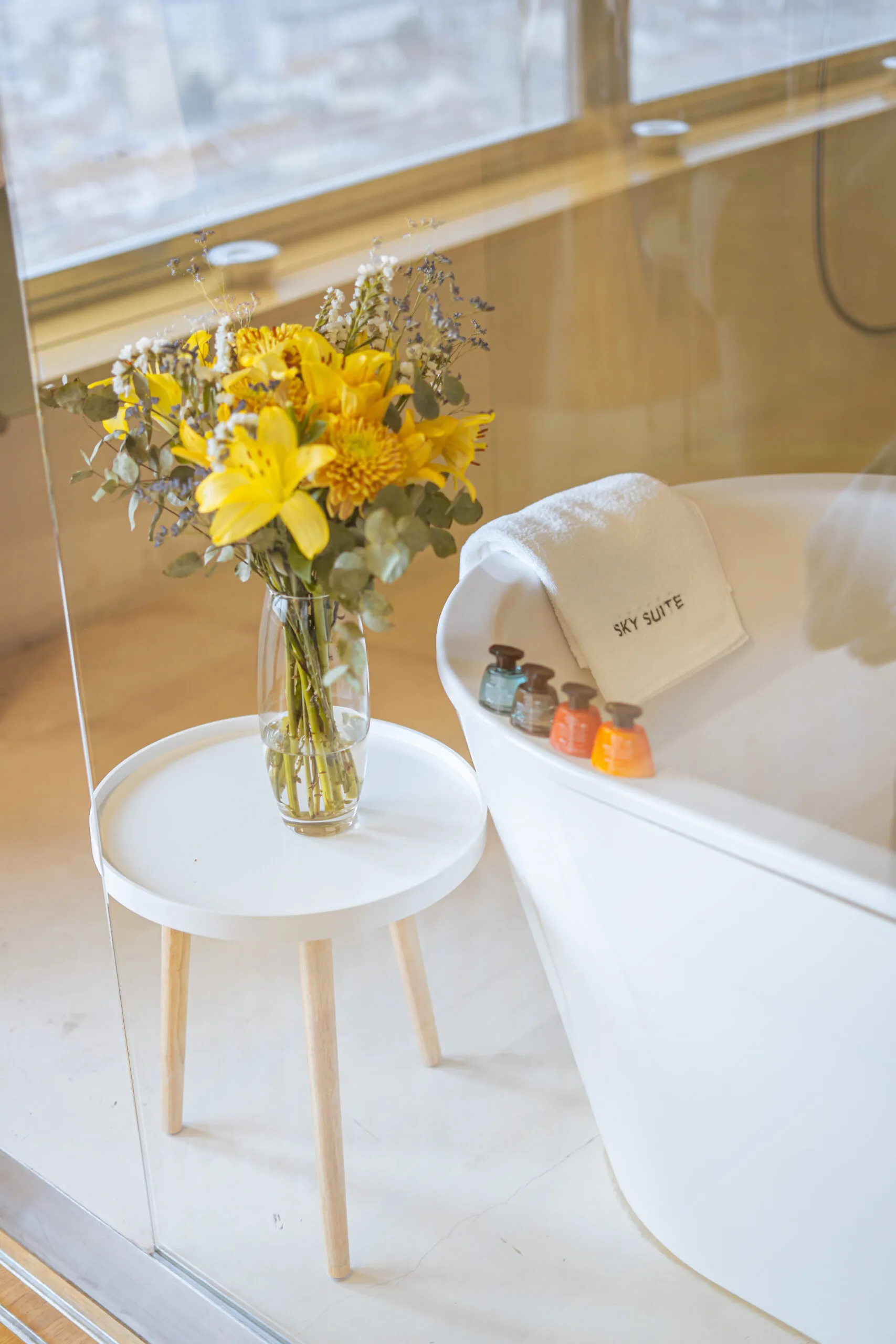 Una bañera blanca con una toalla 'Sky Suite', una mesa pequeña con flores amarillas y una ventana grande con vistas a la ciudad.