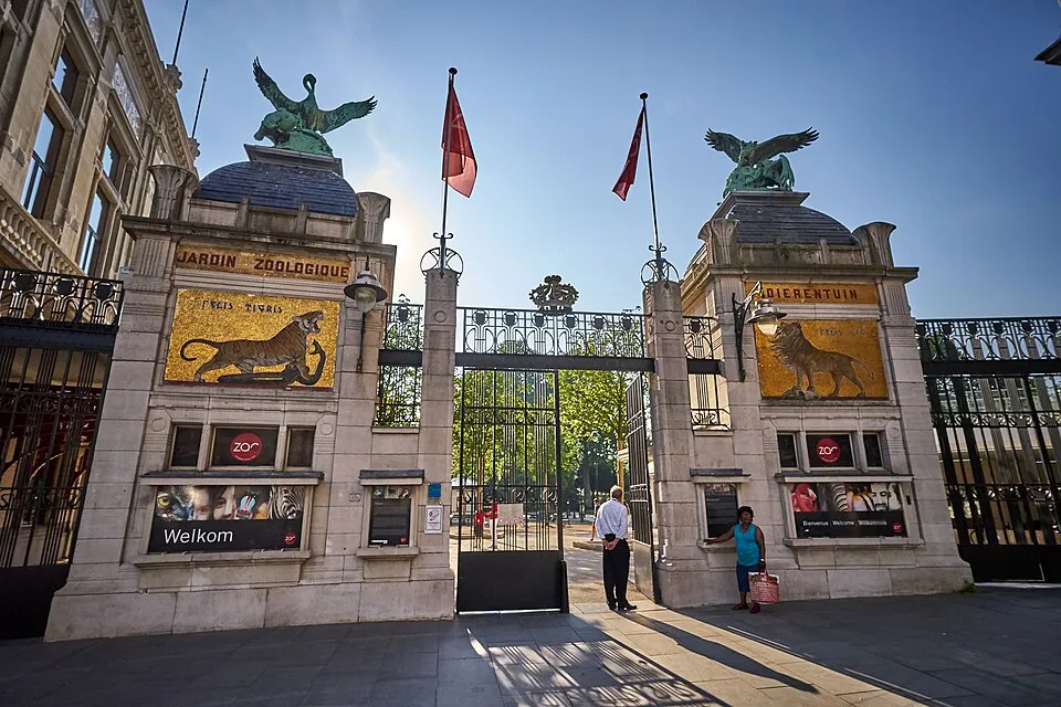 Las torres gemelas de piedra de la entrada del Zoo de Amberes, coronadas por estatuas de águilas de bronce, que presentan paneles de mosaico con leones dorados y puertas de hierro con banderas rojas ondeando bajo un cielo azul.