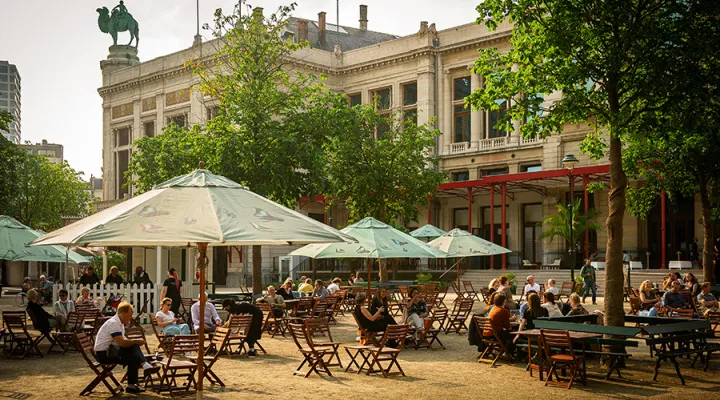 Terraza de café al aire libre con grandes sombrillas verdes y mesas y sillas de madera frente a un gran edificio histórico coronado por una estatua ecuestre en ZOO Antwerpen.