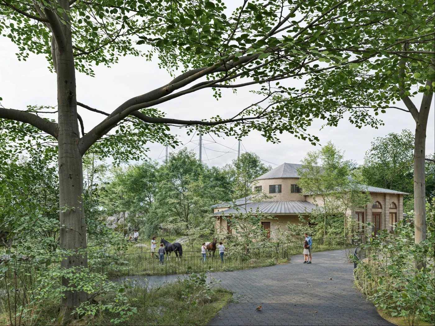 Un sendero serpenteante entre árboles verdes y exuberantes en el Zoo Basel, con visitantes caminando y caballos cerca de un edificio de piedra octogonal