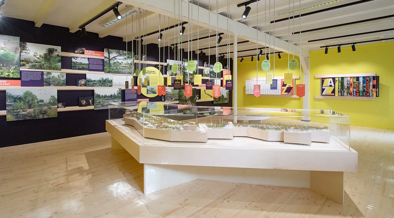 Una toma interior amplia de un espacio de exposición que presenta un modelo a gran escala del Zoo Basel sobre una plataforma blanca, rodeado de vitrinas de cristal, con exhibiciones fotográficas en una pared negra a la izquierda y paneles coloridos colgando del techo.