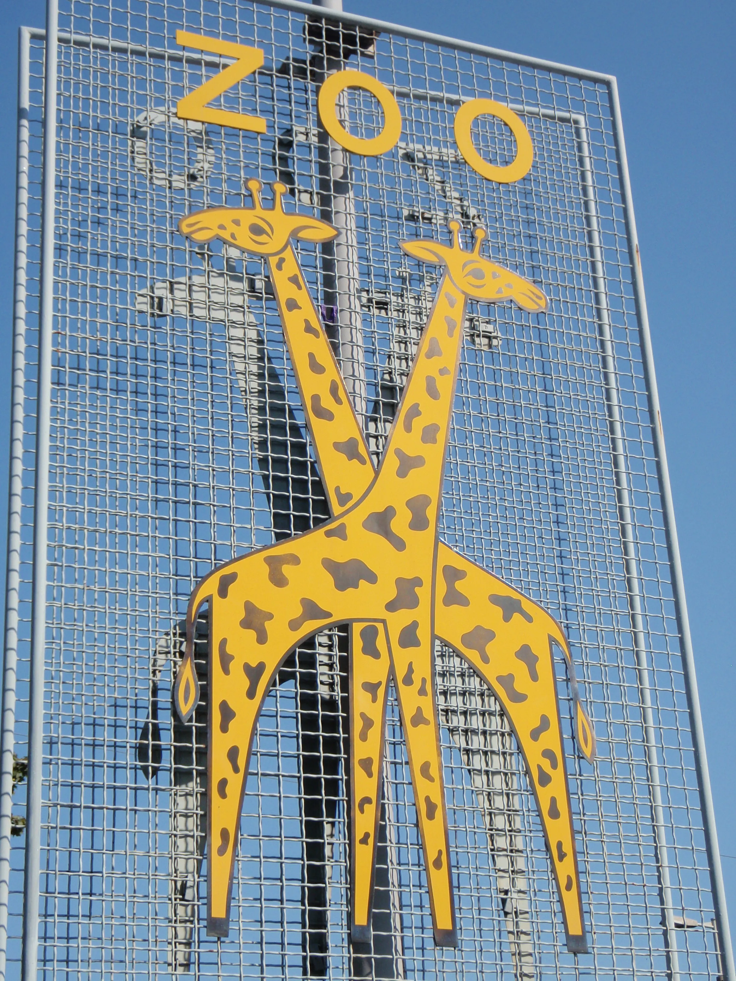Gran letrero metálico exterior con el logotipo del Zoo Basel, que muestra dos jirafas amarillas y la palabra ZOO en letras amarillas sobre un cielo azul.