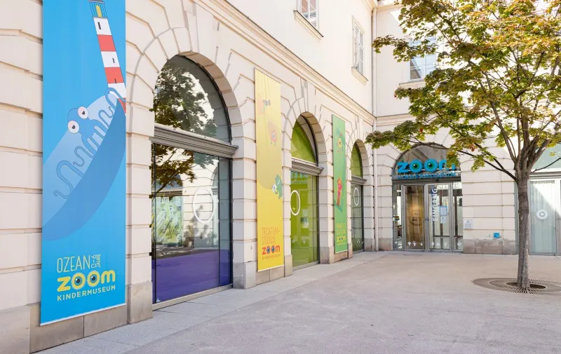 Kolorowe banery z logo ZOOM Children's Museum wiszą na fasadzie budynku z łukowymi oknami i drzewem przed nim.