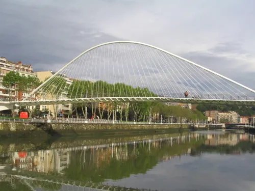 White curved tied-arch pedestrian bridge spanning a river with reflections in the water and trees along the bank.