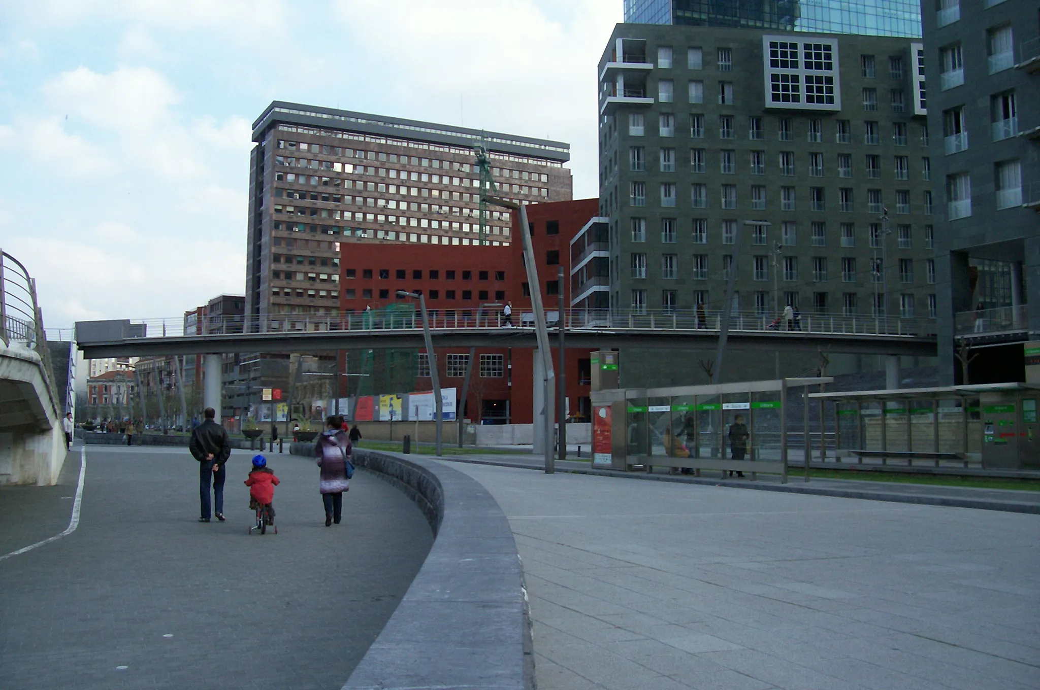 A paved pedestrian walkway curves alongside a white bridge structure, with people walking and modern apartment buildings in the background.