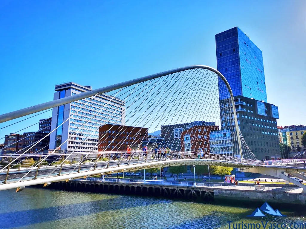 White tied-arch pedestrian bridge spanning a river with cable stays, people walking across, modern glass buildings and blue sky in background