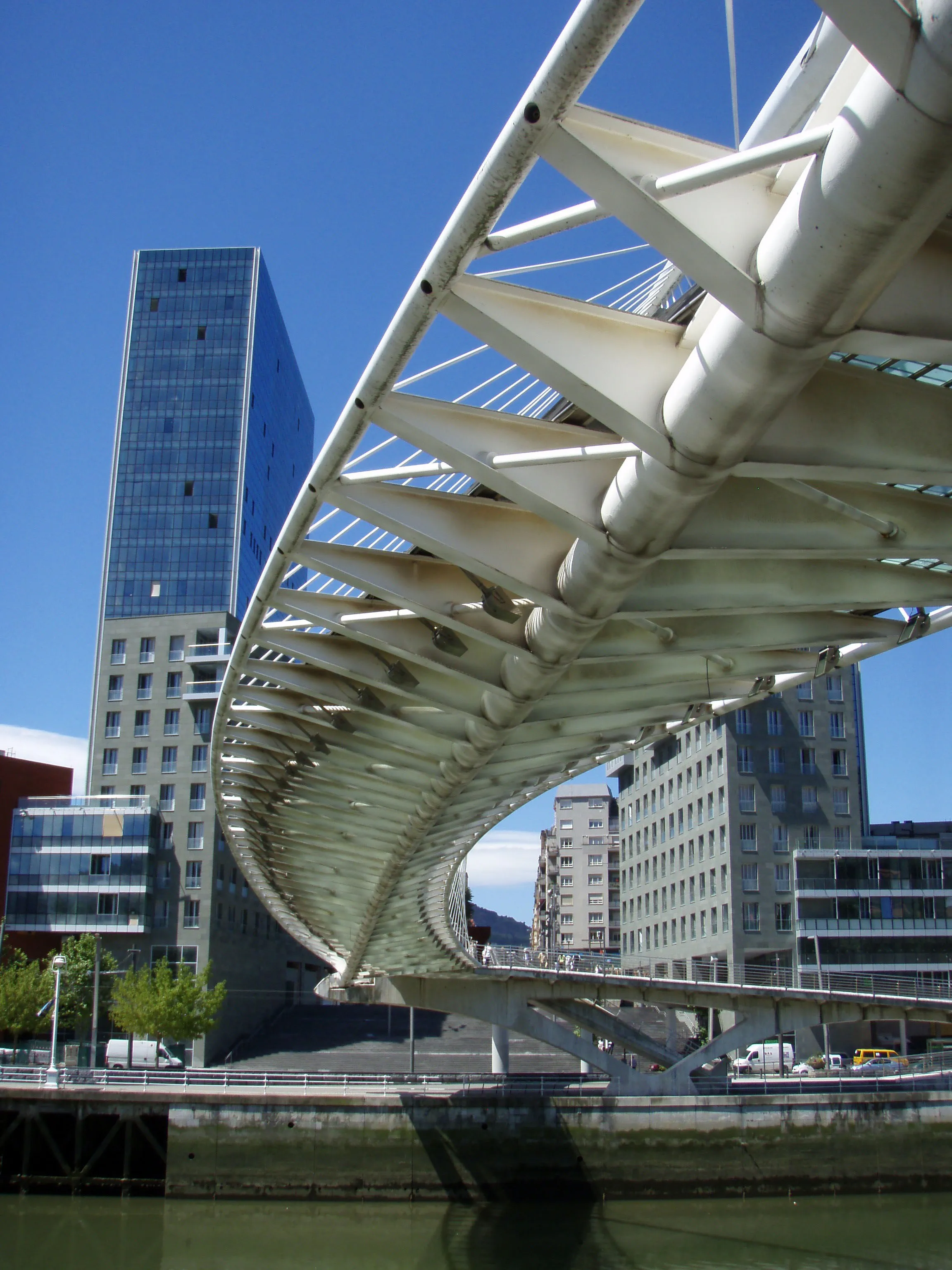 Low-angle view of the white curved steel structure and glass deck of the Zubizuri footbridge, with a tall modern glass building in the background under a clear blue sky.
