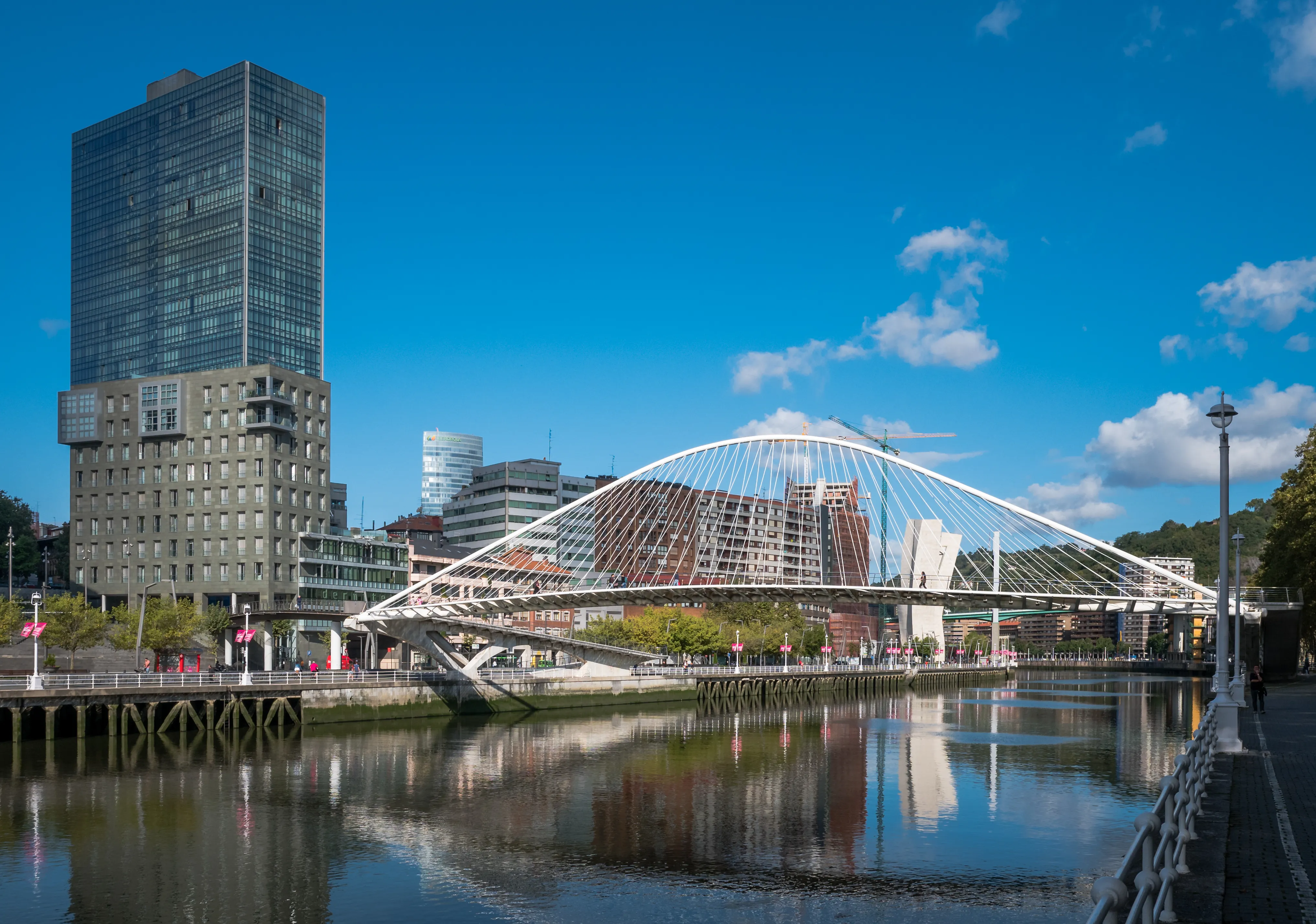 A white tied-arch pedestrian bridge spans a calm river reflecting the structure, with modern high-rise buildings in the background under a blue sky.