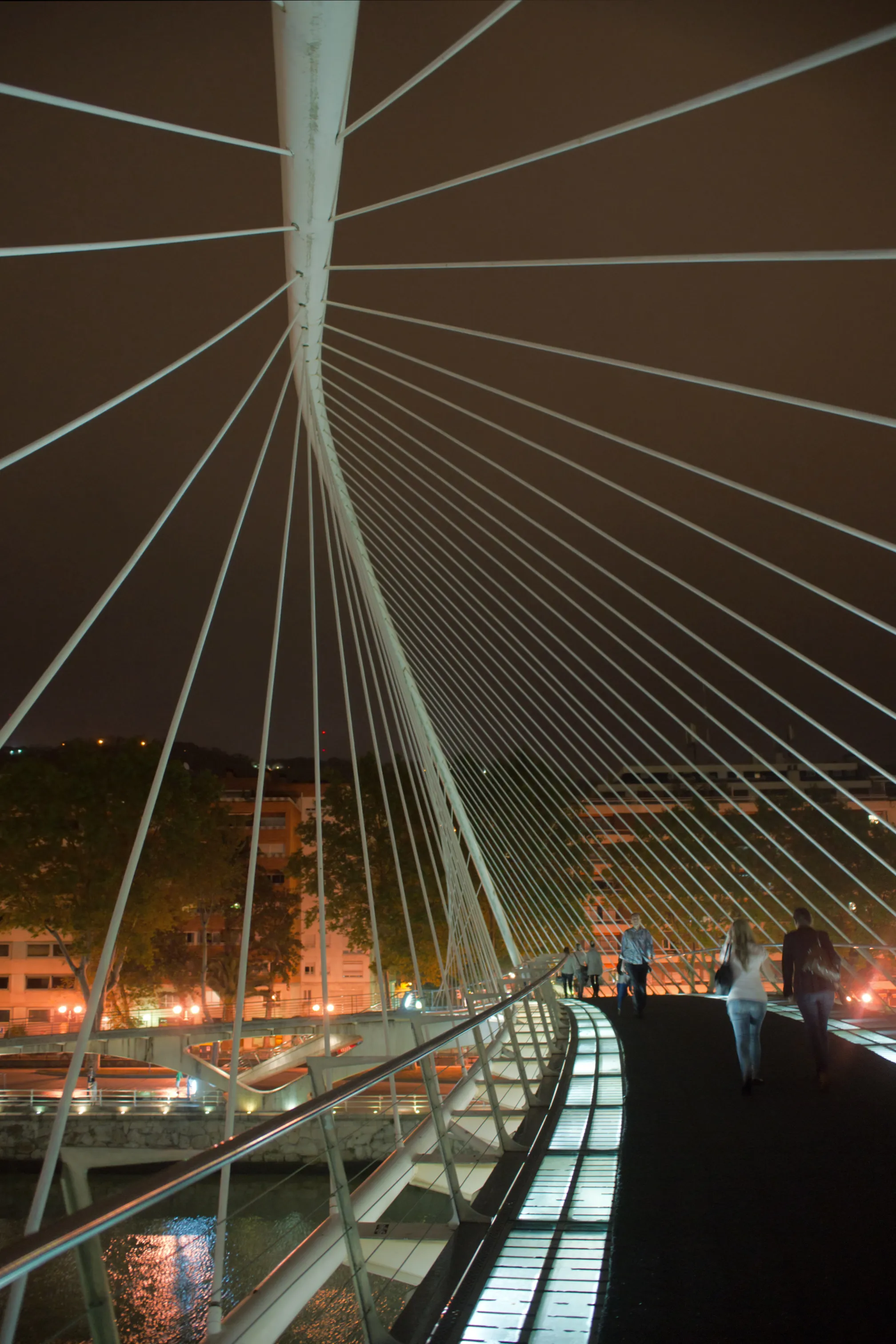 Night view of the Zubizuri footbridge showing its white curved arch, radiating cable stays, and illuminated glass-block walkway with pedestrians crossing over the Bilbao Estuary.